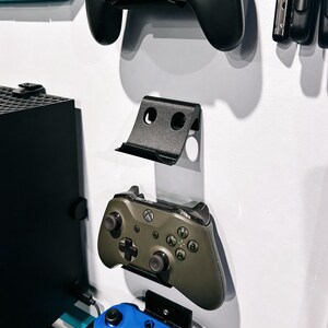 Könnte beinhalten: Eine vertikale, an der Wand montierte schwarze Kunststoffhalterung für Gaming-Controller mit geschwungenem Design. Die Halterung zeigt einen schwarzen Nintendo Switch Pro Controller, einen olivgrünen Xbox Controller und einen blauen Xbox Controller.
