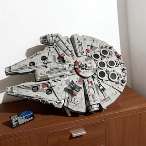 Può includere: Un modello dettagliato dell'astronave Millennium Falcon, principalmente grigio con accenti rossi e neri. Il modello è su una superficie di legno, con una piccola carta accanto. L'astronave presenta dettagli intricati e molteplici elementi circolari.