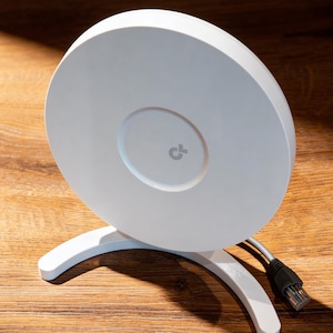 Può includere: Un punto di accesso wireless bianco e circolare con un supporto curvo. Il dispositivo ha un'area centrale incassata con un piccolo logo. Un cavo grigio con un connettore nero si estende dal dispositivo. Il punto di accesso è su una superficie di legno.