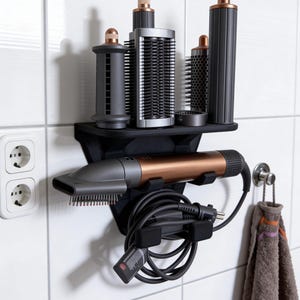 Può includere: Un organizer nero da parete per strumenti per lo styling dei capelli con un ripiano. Contiene un asciugacapelli color rame e nero e diversi accessori per lo styling. L'organizer è montato su una parete piastrellata bianca. Un asciugamano è appeso a un gancio a destra.