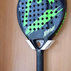 Könnte beinhalten: Schwarzer und grüner Xenon Padel-Schläger mit schwarzem Griff. Der Schläger hat eine Reihe von Löchern und grüne Akzente. Der Schläger ist auf einem weißen Halter vor einem Holzgrund befestigt.