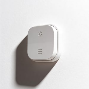 Op de afbeelding: Een witte, vierkante wandschakelaar met afgeronde randen. De schakelaar heeft een sneeuwvloksymbool aan de bovenkant en drie horizontale lijnen aan de onderkant. Hij is op een witte muur gemonteerd en werpt een schaduw.