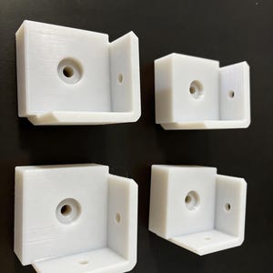 Op de afbeelding: Vier witte, L-vormige kunststof hoekbeugels. Elke hoekbeugel heeft een vierkante basis met een centraal gat en een loodrechte zijde met een kleiner gat. De beugels zijn ontworpen voor montage in een hoek van 90 graden.