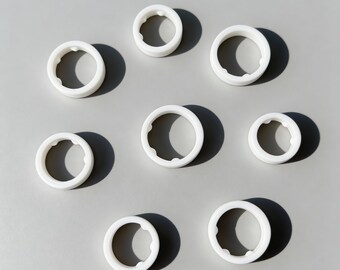 May include: A collection of eight white, ring-shaped objects arranged on a light gray surface. Each ring has a rounded outer edge and a textured inner edge. The objects cast dark shadows, creating a sense of depth.