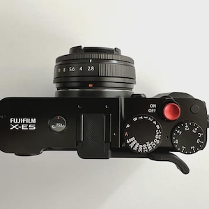 Könnte beinhalten: Schwarze Fujifilm X-E5 Kamera mit Objektiv. Die Kamera hat mehrere Einstellräder und Knöpfe, darunter einen Ein-/Ausschalter und einen roten Knopf. Der Text "FUJIFILM X-E5" ist auf dem Kameragehäuse zu sehen.