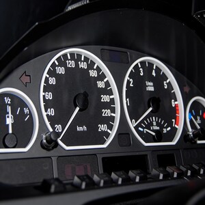 May include: A close-up of a black car dashboard featuring white-faced gauges. The speedometer displays up to 240 km/h. Other gauges show fuel level and RPM. The dashboard includes buttons and a digital display.