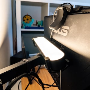 Könnte beinhalten: Eine schwarze Monitor-Lichtleiste, die an der Oberkante eines schwarzen Computermonitors befestigt ist. Die Lichtleiste verfügt über ein langes, rechteckiges, beleuchtetes weißes Panel, das nach vorne gerichtet ist, sowie ein schwarzes Stromkabel.