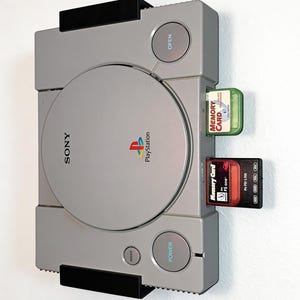 Könnte beinhalten: Eine graue Sony PlayStation-Konsole, die an einer Wand montiert ist, mit kreisförmiger Disc-Abdeckung, Power-Taste, Open-Taste und Reset-Taste. In den vorderen Steckplätzen befinden sich zwei Speicherkarten: eine durchsichtig grüne und eine schwarze.