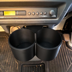 Könnte beinhalten: Ein schwarzer Doppel-Getränkehalter, der unter einem Autoradio am Armaturenbrett eines Fahrzeugs montiert ist. Das Radio verfügt über ein digitales Display, fünf runde Tasten, einen Power-Knopf und den Schriftzug VTUNE A. Der Halter hat zwei Fächer.