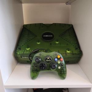 Op de afbeelding: Een groene, doorschijnende Xbox-console en bijbehorende controller. De console is rechthoekig met het Xbox-logo. De controller heeft gekleurde knoppen en een D-pad. De items worden op een witte plank gepresenteerd.