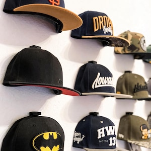Könnte beinhalten: Eine Sammlung von Baseballkappen, die an einer Wand befestigt sind, mit verschiedenen Designs wie einem San Francisco Giants-Logo, einem Batman-Emblem und Texten wie "Drop", "Iowa" und "HW Motor 13". Die Kappen haben verschiedene Farben.