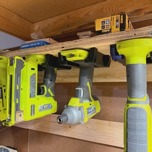Könnte beinhalten: Drei lindgrüne und graue Elektrowerkzeuge hängen an schwarzen Kunststoffhalterungen an einem Holzregal. Die Werkzeuge umfassen eine Stichsäge, einen Nagler und einen Schlagschrauber. Eine gelbe DeWalt-Box steht auf dem Regal über den Werkzeugen.
