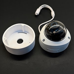 Könnte beinhalten: Eine weiße Dome-Überwachungskamera mit einer klaren, abgerundeten Linse und einem weißen Sockel. Die Kamera wird neben ihrer weißen Montagebasis und einem weißen Kabel mit einem Stecker gezeigt. Der Hintergrund ist schwarz.