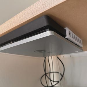 Könnte beinhalten: Eine schwarze und silberne Sony PlayStation 4 Spielkonsole, die mit einem weißen Kunststoffhalterungssystem unter einer Holzoberfläche montiert ist. Die Konsole ist horizontal angebracht, mit sichtbaren schwarzen Kabeln an der Rückseite.