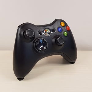 Könnte beinhalten: Schwarzer Xbox 360-Controller mit farbigen Tasten und zwei Analogsticks. Der Controller hat ein Steuerkreuz, eine zentrale Xbox-Taste und Start/Zurück-Tasten. Der Controller liegt auf einer hellen Oberfläche.