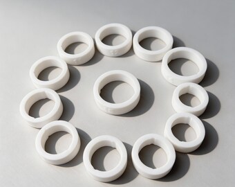 May include: A collection of white, circular objects arranged in a circular pattern. Each ring has a smooth, matte finish and a consistent thickness. The objects are evenly spaced against a light gray background, creating a minimalist aesthetic.
