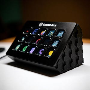 Könnte beinhalten: Eine schwarze Stream Deck Steuerungskonsole mit fünfzehn quadratischen Tasten und beleuchteten Symbolen. Das Gerät hat eine abgeschrägte Oberfläche und ein Seitenteil mit sechseckigem Muster. Der Markenname Stream Deck ist oben weiß aufgedruckt.