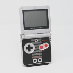 Könnte beinhalten: Eine silberne und schwarze Game Boy Advance SP Handheld-Spielkonsole mit Klappdesign. Das Gerät verfügt über einen Bildschirm mit dem Text Game Boy Advance SP, ein schwarzes Bedienfeld mit Steuerkreuz, zwei roten A- und B-Tasten und Nintendo-Logo.