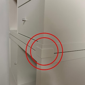 Könnte beinhalten: Ein weißer Kunststoffeckenschutz, der an der Kante eines weißen Regals angebracht ist. Der Schutz ist L-förmig und passt über die Ecke, an der zwei Möbelteile aufeinandertreffen. Das Regal verfügt über Schubladen mit kleinen silbernen Knöpfen.
