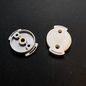 Op de afbeelding: Twee ronde onderdelen, één lichtgrijs en één wit, voorzien van een centraal messingkleurig schroefdraadinzetstuk. Het grijze onderdeel toont een verzonken onderkant met twee montagegaten, terwijl het witte onderdeel een textuur heeft.