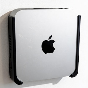 Könnte beinhalten: Ein silberner Apple Mac Mini Computer, der in einer schwarzen Wandhalterung befestigt ist. Das Gerät hat eine quadratische Form mit abgerundeten Ecken und ein markantes schwarzes Apple-Logo in der Mitte der Oberseite sowie sichtbare Anschlüsse.