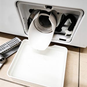 Könnte beinhalten: Eine weiße Kunststoff-Ablaufrinne und eine rechteckige weiße Auffangschale unter der geöffneten Wartungsklappe einer Waschmaschine. Ein schwarzer gerippter Schlauch ist im Inneren des Maschinengehäuses neben der runden Ablauföffnung sichtbar.