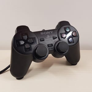 Könnte beinhalten: Schwarzer PlayStation 3-Controller mit Sony-Logo. Der Controller hat ein Steuerkreuz, Analog-Sticks und Aktionstasten. Der Controller ist mit einem schwarzen Kabel verbunden.