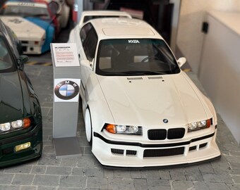 May include: A white BMW sports car with black accents and the text "KYZA" on the windshield. The car is parked next to a dark green BMW. A BMW logo is on a display stand.