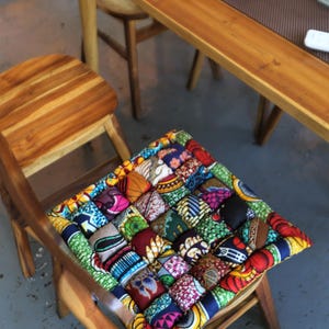 May include: A square cushion with a patchwork design, featuring a variety of colorful African fabric patterns. The cushion is placed on a wooden chair, showcasing a blend of textures and vibrant hues. The chair and cushion are in a room with wooden tables.