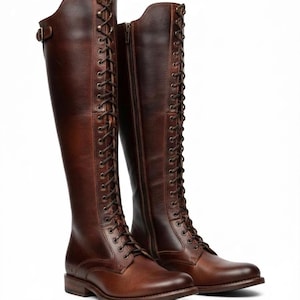 May include: A pair of knee-high brown leather boots featuring a full-length front lace-up design with brass-toned eyelets, a side zip closure, and a small adjustable buckle strap at the top of the shaft, set on a low stacked heel.