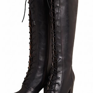 May include: A pair of black knee-high boots featuring a full-length front lace-up closure with black laces, a side zip, a rounded toe, and a sturdy block heel. The boots have a smooth finish and reach just below the knee with a structured shaft.