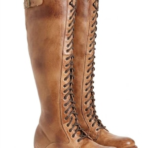 May include: A pair of knee-high tan leather boots featuring a full-length front lace-up design with dark brown laces, a rounded toe, a low stacked heel, and a small adjustable buckle strap detail located at the top outer side of the boot shaft.