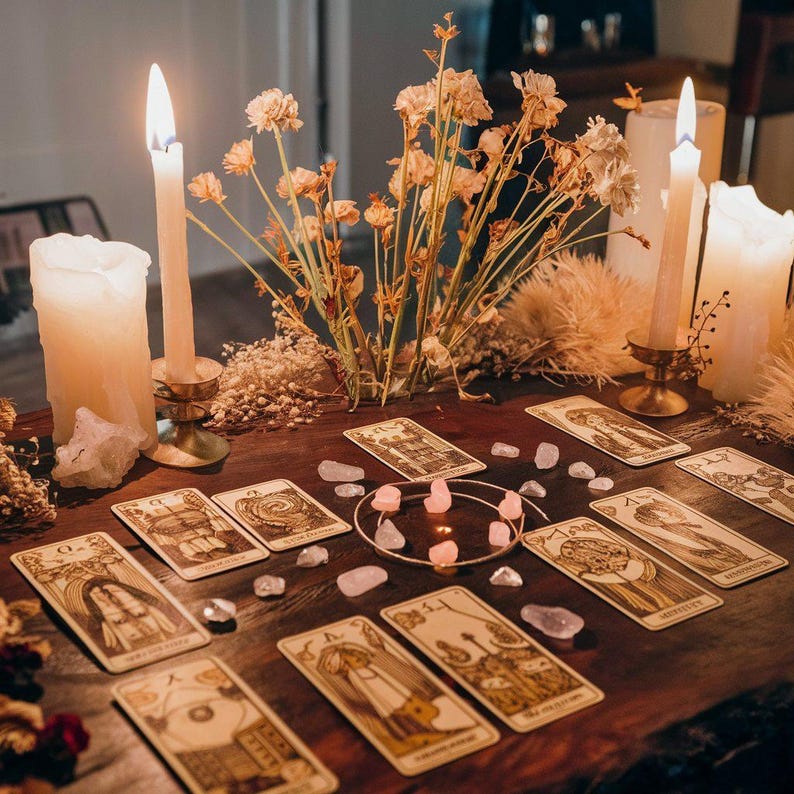 Puede incluir: Una mesa de madera exhibe cartas del tarot, cristales y velas encendidas. Las cartas presentan ilustraciones detalladas en tonos marr&oacute;n y beige. Flores secas y velas adicionales crean un ambiente m&iacute;stico. Cristales de cuarzo rosa est&aacute;n dispersos.