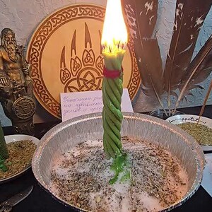 May include: A lit green candle with a twisted design stands in a metal bowl filled with herbs and salt. A second green candle is lit in a small dish. A wooden plaque with a bear claw design and a statue are in the background.