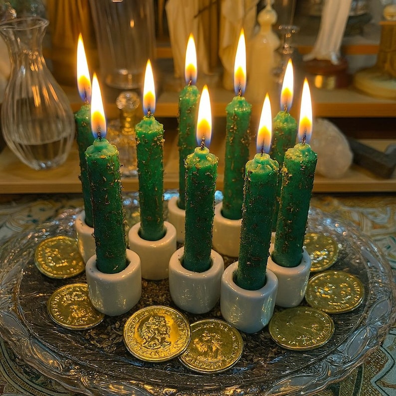 Puede incluir: Un conjunto de velas verdes encendidas, dispuestas en soportes de cer&aacute;mica blanca sobre un plato de cristal. Varias monedas de oro est&aacute;n dispersas alrededor de las velas, creando un ambiente m&iacute;stico.