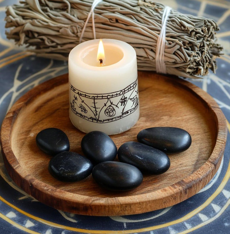 May include: A lit cream-colored candle with a black design sits on a wooden tray with black stones. A bundle of dried sage is behind the candle. The candle has a black design around the middle.