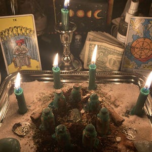 Puede incluir: Una bandeja plateada contiene un arreglo espiritual con velas verdes encendidas, peque&ntilde;as botellas verdes y una dispersi&oacute;n de hierbas y cristales. Se ven cartas del tarot y moneda estadounidense, creando un ambiente m&iacute;stico.