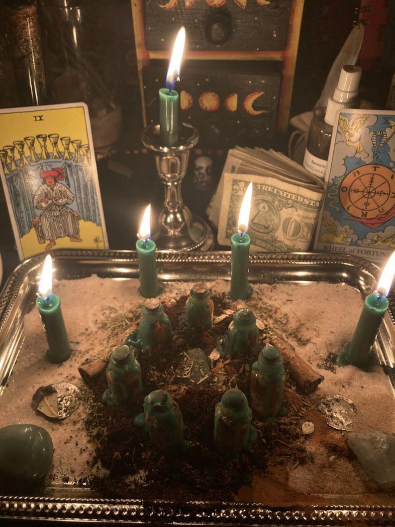 Puede incluir: Una bandeja plateada contiene un arreglo espiritual con velas verdes encendidas, peque&ntilde;as botellas verdes y una dispersi&oacute;n de hierbas y cristales. Se ven cartas del tarot y moneda estadounidense, creando un ambiente m&iacute;stico.