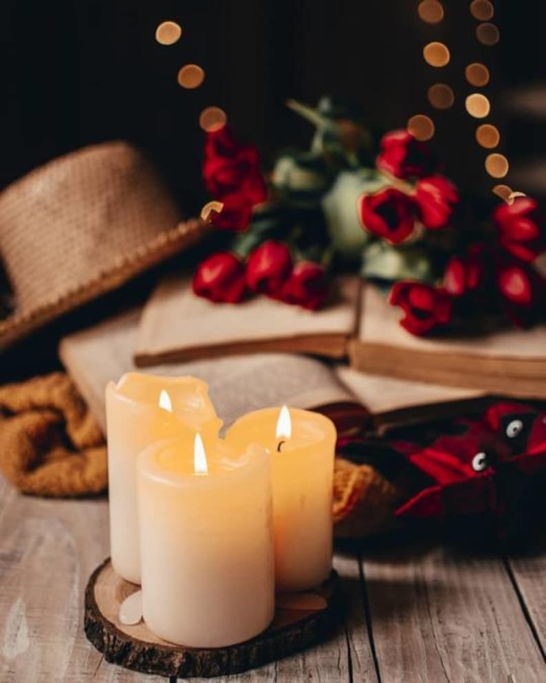 May include: Three lit ivory-colored pillar candles on a wooden slice, with a backdrop of open books, red tulips, and a straw hat. The scene evokes a cozy, warm ambiance, perfect for relaxation and reading.