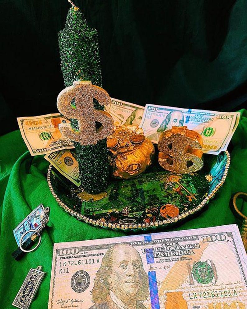 Puede incluir: Una vela verde cubierta de purpurina con un signo de d&oacute;lar dorado y una bandeja decorativa llena de billetes de 100 d&oacute;lares estadounidenses y monedas de oro. Tambi&eacute;n hay un llavero con un billete de 100 d&oacute;lares en miniatura y un colgante con la palabra "ONE".