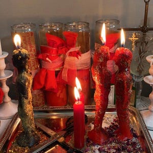 Pu&ograve; includere: Una collezione di candele accese di varie forme e dimensioni, tra cui candele rosse a forma di figure e una candela nera alta. Le candele sono su un vassoio d'argento con forbici e fiori secchi. Barattoli con nastri rossi sullo sfondo.