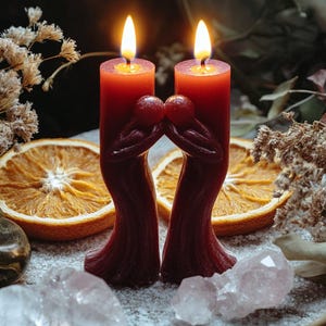 May include: Two lit, burgundy-colored pillar candles with a sculpted design of two figures embracing. The candles are set against a backdrop of dried orange slices, crystals, and dried flowers, creating a warm, inviting ambiance.