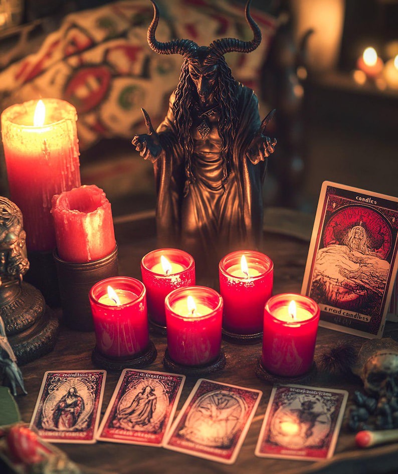 May include: A bronze statue of a horned figure stands behind several lit red candles in glass holders. Tarot cards with illustrations are arranged in front of the candles. A large red candle and a smaller red candle are also lit.