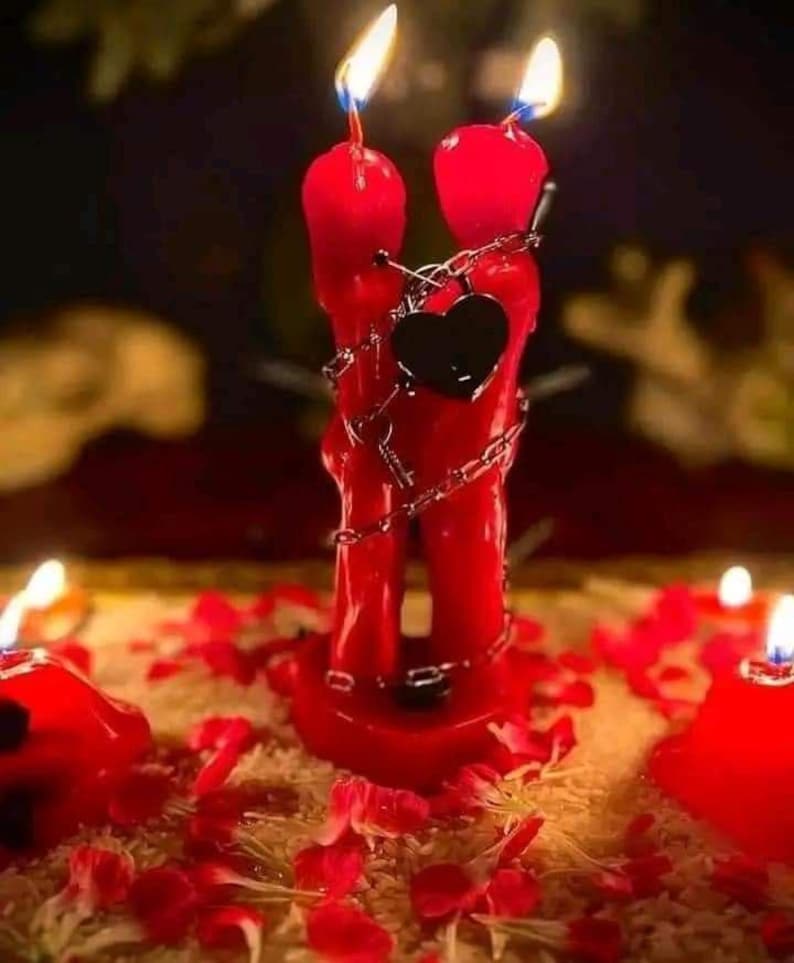 May include: A pair of red candles, shaped to resemble two figures embracing, with flames burning at the top. A black heart pendant hangs between them, and they are wrapped in a silver chain. The candles are surrounded by rose petals.
