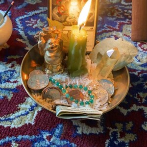 Puede incluir: Un cuenco dorado contiene una estatua de Buda sonriente, monedas, arroz y un collar de cuentas verdes. Una vela verde encendida y una tarjeta con el texto "Lucky Offering" tambi&eacute;n est&aacute;n presentes. Sobre una alfombra estampada.