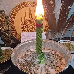 Puede incluir: Una vela verde encendida con un dise&ntilde;o retorcido se encuentra en un cuenco de metal lleno de hierbas y sal. Una segunda vela verde est&aacute; encendida en un plato peque&ntilde;o. Una placa de madera con un dise&ntilde;o de garra de oso y una estatua est&aacute;n en el fondo.