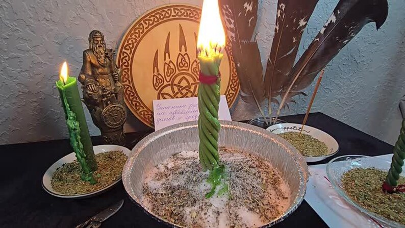 Puede incluir: Una vela verde encendida con un dise&ntilde;o retorcido se encuentra en un cuenco de metal lleno de hierbas y sal. Una segunda vela verde est&aacute; encendida en un plato peque&ntilde;o. Una placa de madera con un dise&ntilde;o de garra de oso y una estatua est&aacute;n en el fondo.