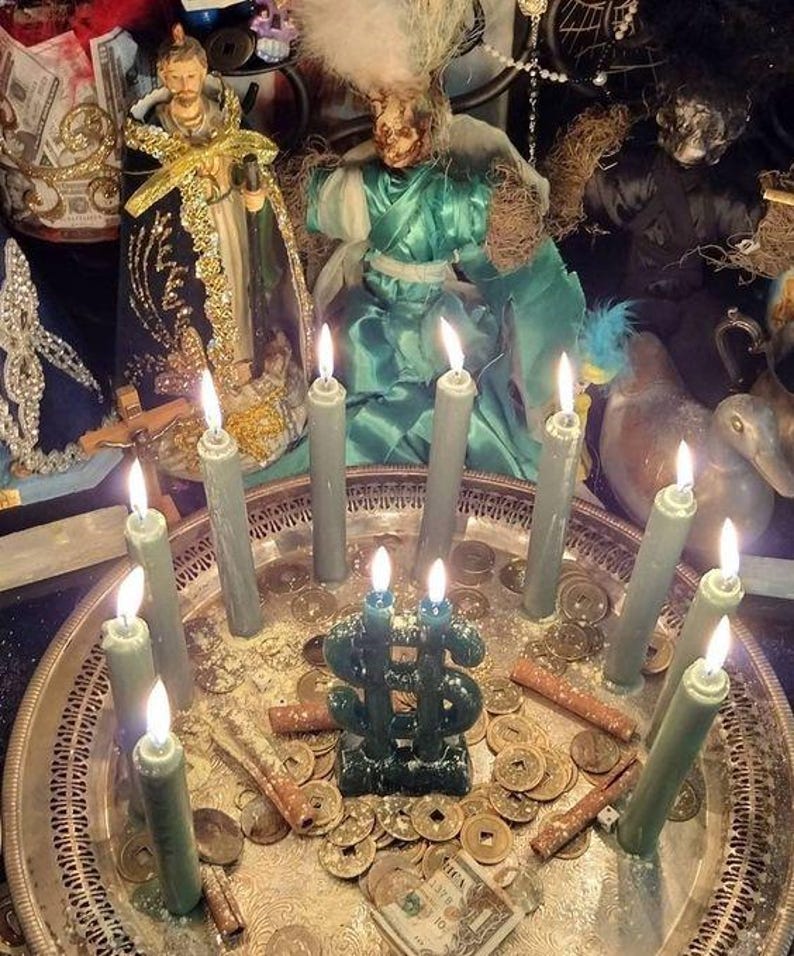 May include: A silver tray holds lit green candles, coins, and a green candle shaped like a dollar sign. Two religious figurines are in the background. The scene is illuminated by the candle flames.