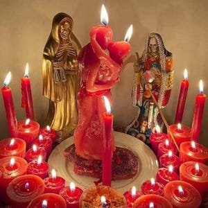 May include: A spiritual altar with lit red candles and statues. A central red candle depicts two figures embracing. Two gold statues and a third statue with a skeletal face are also present. The scene is illuminated by the candle flames.