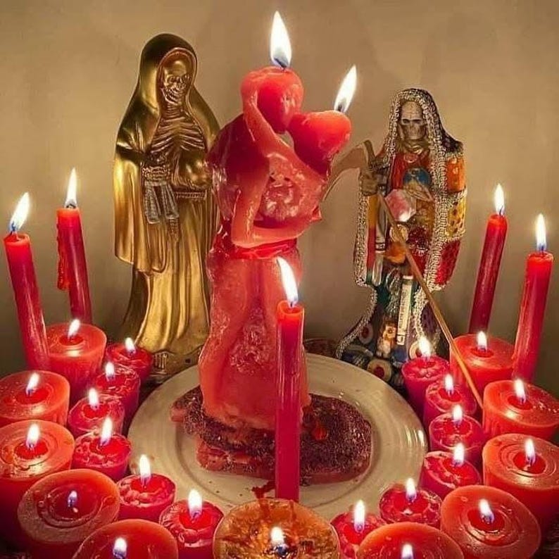 May include: A spiritual altar with lit red candles and statues. A central red candle depicts two figures embracing. Two gold statues and a third statue with a skeletal face are also present. The scene is illuminated by the candle flames.